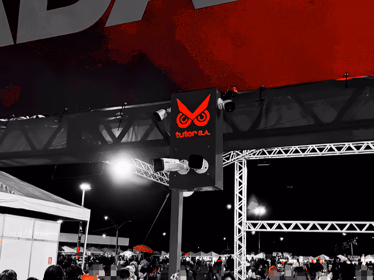 Totem de vigilância com câmeras de segurança em evento noturno, com logotipo vermelho iluminado e público ao fundo.